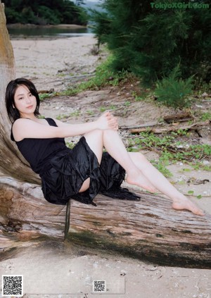 A woman in a black dress sitting on a log on the beach.