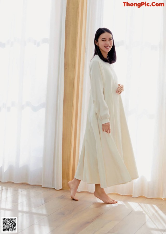 A woman standing in front of a window wearing a white dress.