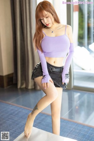 A woman in a purple top and black skirt posing by a window.