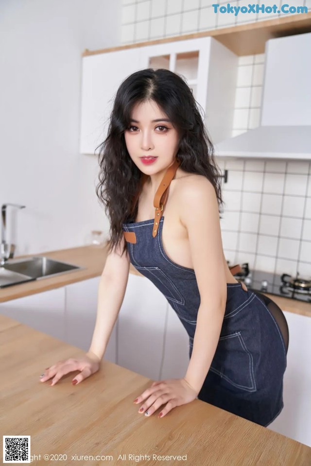 A woman in a denim dress leaning on a kitchen counter.